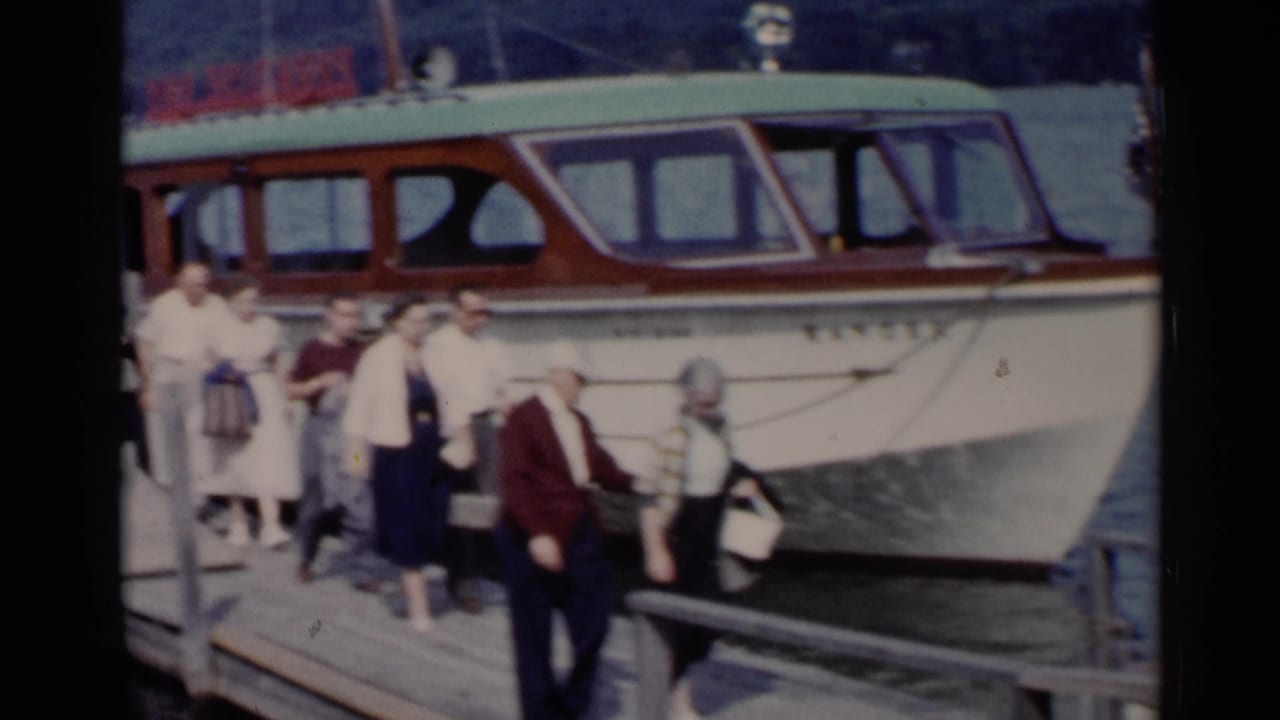 1955 Lake George home-movie frame showing passengers boarding near a white and brown motorboat at a wooden dock.