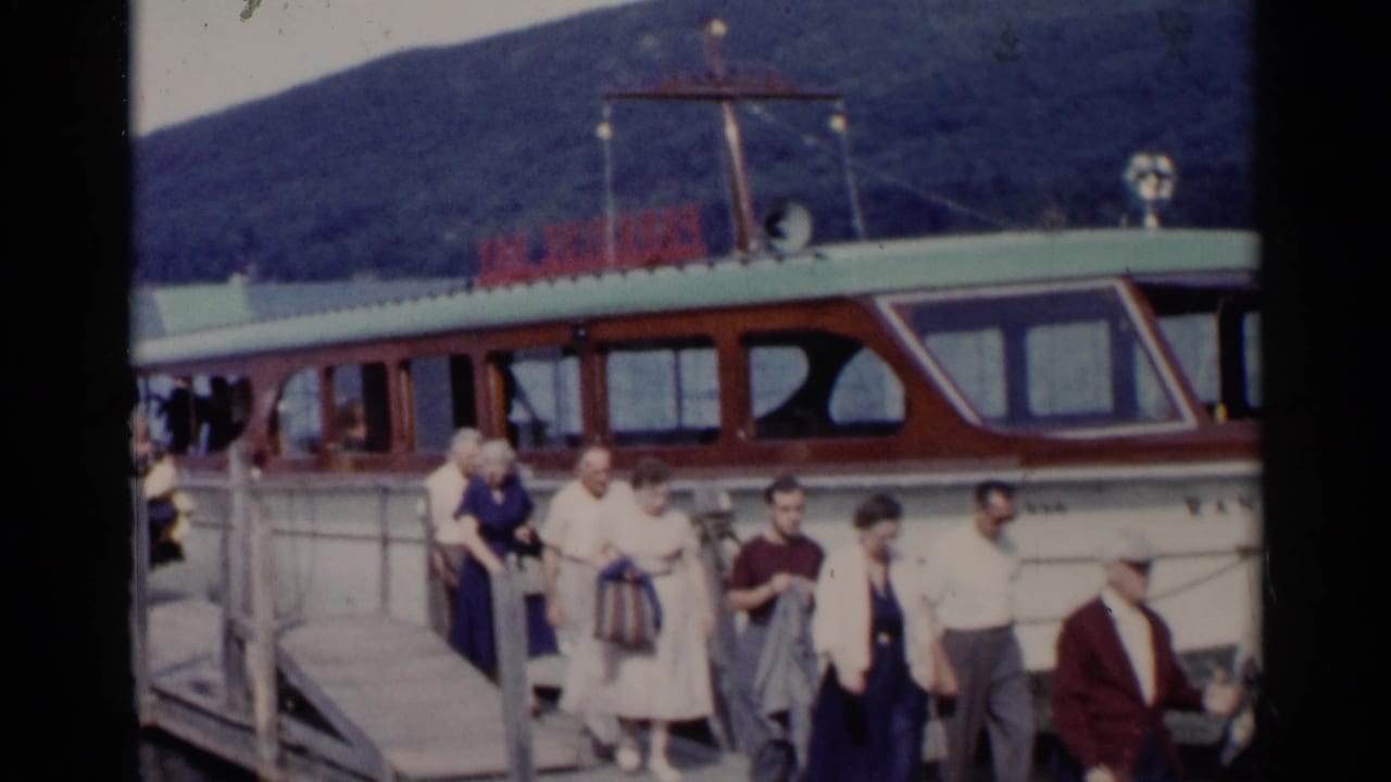 1955 Lake George frame with adults in summer attire moving along a dock beside a cabin motorboat and wooded hills.