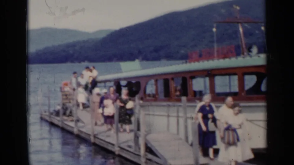 Wide 1955 Lake George view of passengers on a wooden dock beside a motorboat, with lake water and wooded hills behind.