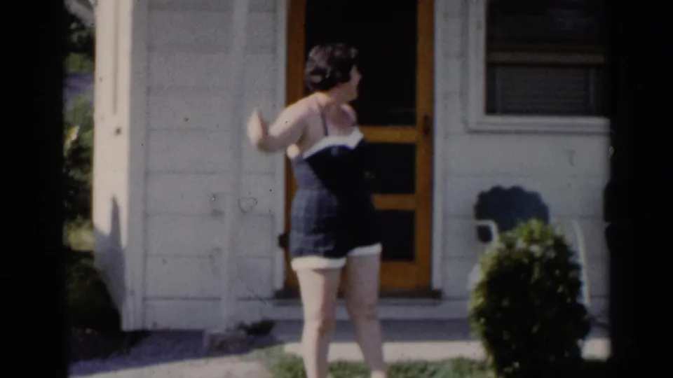 1955 home-movie frame showing a woman in a dark swimsuit outside a light-colored building near a doorway.