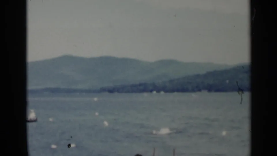 Lake George home-movie frame with open water, distant tree-covered shoreline, hazy hills, and small distant boating activity.