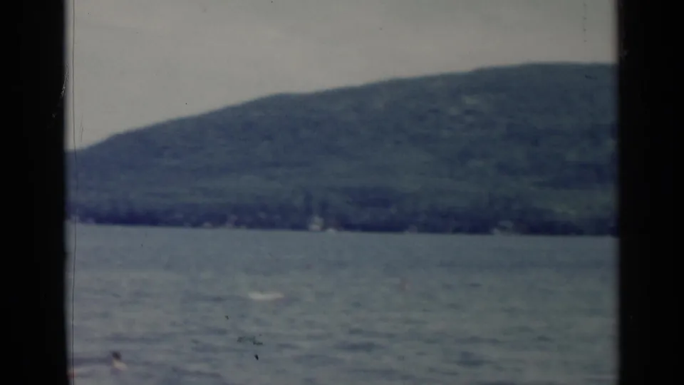 1955 Lake George frame showing open water, a distant wooded hill, and small people or swimmers near the bottom edge.