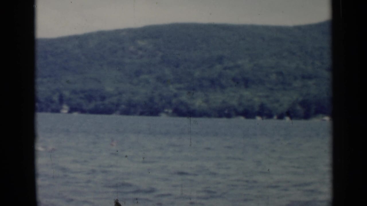 1955 Lake George home-movie view of lake water, wooded hillside, pale sky, and dark vertical film-frame edges.