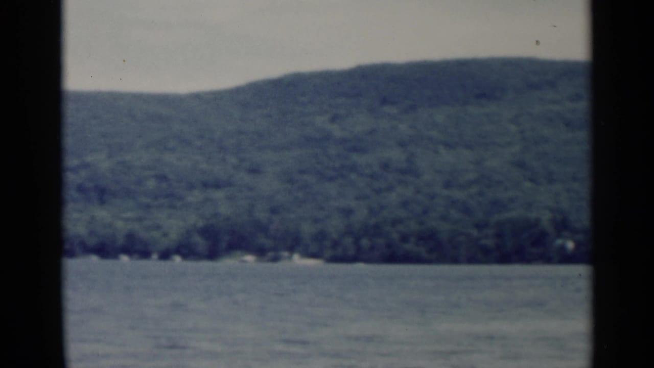 Lake George shoreline and wooded hill in soft 1950s 8mm color, with water in the foreground and dark frame edge visible.