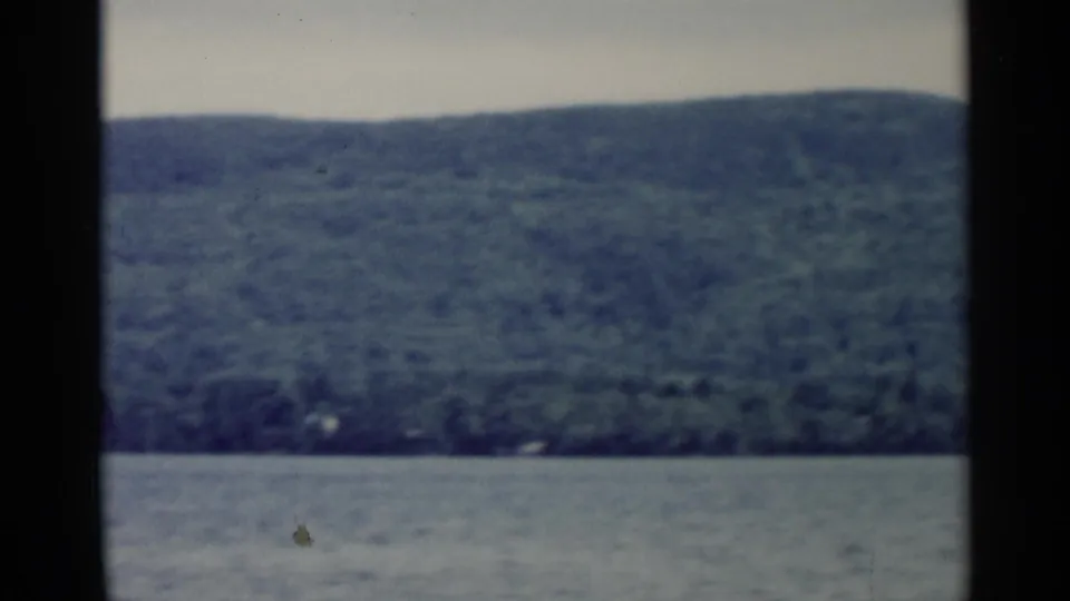 Soft 8mm frame of Lake George water below a broad green wooded hillside, with a distant shore near the horizon.