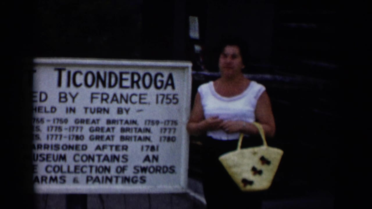 Woman stands beside a Fort Ticonderoga sign while holding a pale tote bag.