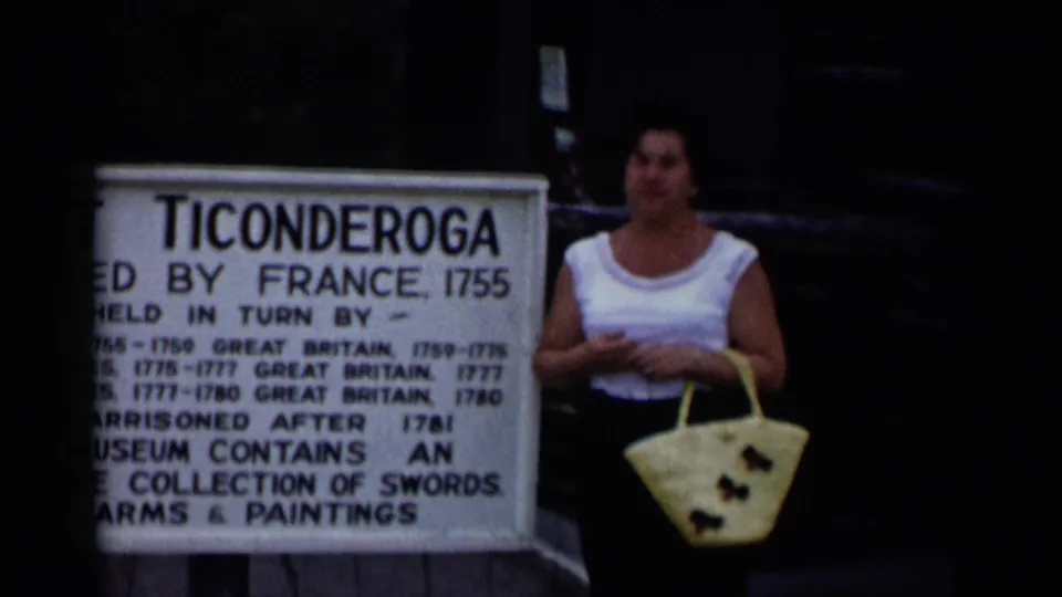 In 1955 Lake George-area home-movie footage, a woman poses beside a Fort Ticonderoga information sign with a pale tote bag.