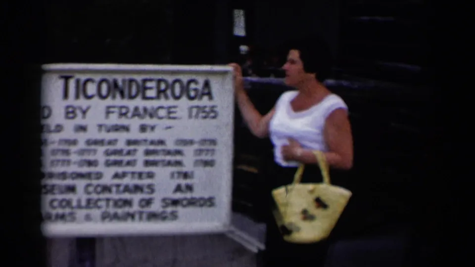 A woman in a white sleeveless top gestures beside the Fort Ticonderoga sign during a 1955 Lake George-area tourist stop.