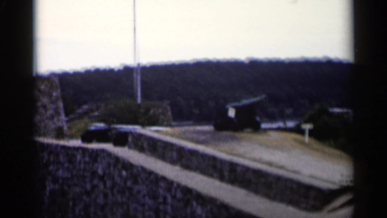 A handheld 1955 view of Fort Ticonderoga-area grounds shows a cannon, stone walls, paved walkway, flagpole, and wooded hills.