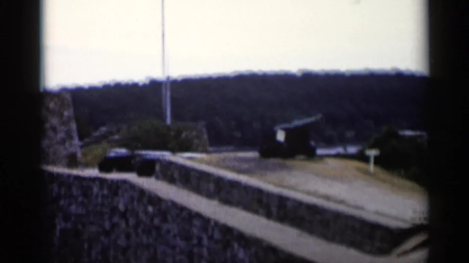 A handheld 1955 view of Fort Ticonderoga-area grounds shows a cannon, stone walls, paved walkway, flagpole, and wooded hills.