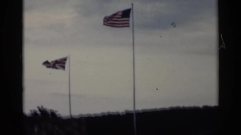 Two flags, including American and British flags, wave on tall poles over dark treetops in the 1955 Lake George-area footage.