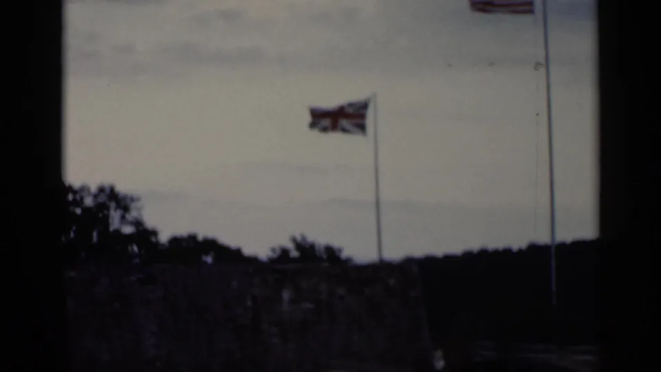 A British flag waves on a tall flagpole above trees and low stonework in the Lake George-area historic-site scene.