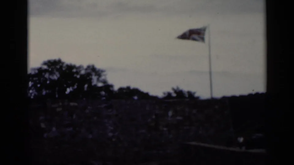 1955 Lake George home-movie frame showing a stone wall, trees, cloudy sky, and a flag waving from a tall pole.