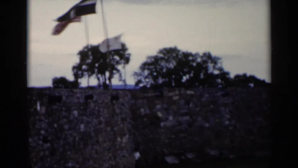 1955 Lake George frame with two visible flags flying over a rough stone wall, with trees and sky in the background.
