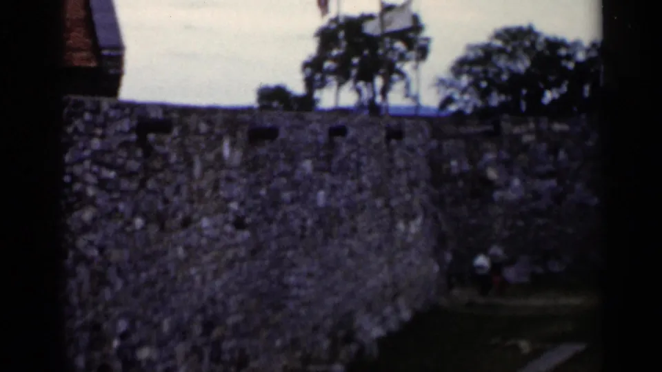 1955 Lake George frame showing a close section of stone wall, trees, grass, and small distant people near the grounds.