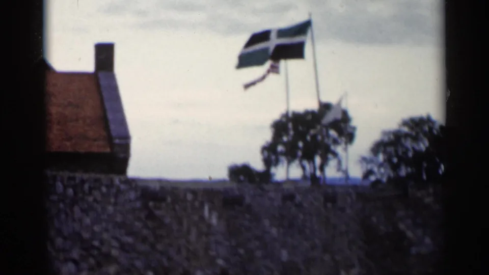 1955 Lake George home-movie frame with three flags above a stone wall, plus roofline, trees, and cloudy sky.