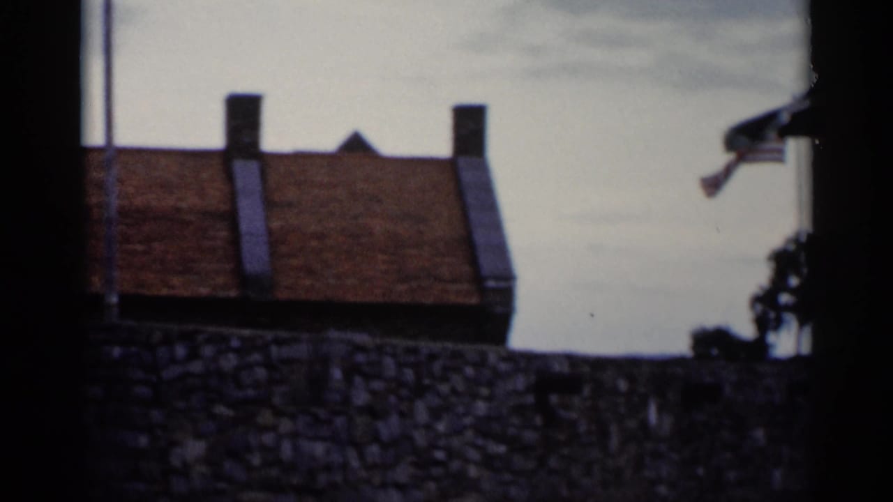 1955 Lake George home-movie frame showing a stone wall, red-tiled roof, chimneys, pale sky, and dark edge framing.