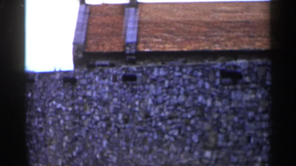 1955 Lake George frame with a broad stone wall, red roof tiles, small wall openings, and a bright film-edge flare at left.