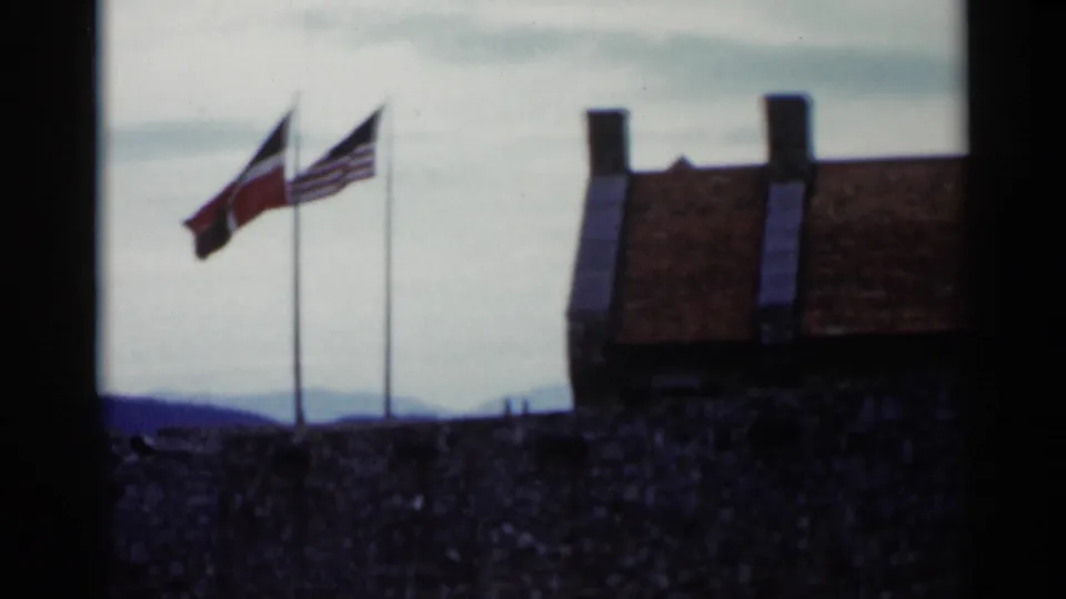 1955 Lake George home-movie frame showing two flags, including an American flag, beside a stone wall, roofline, sky, and distant hills.