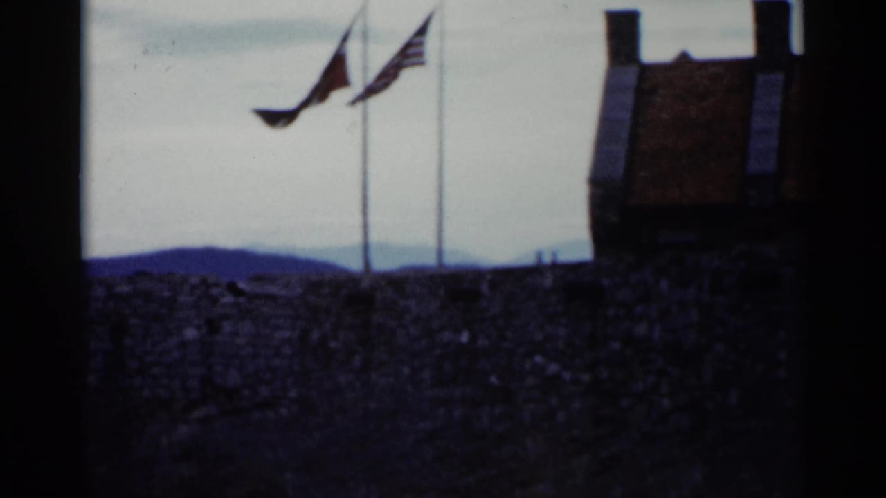Two flags on poles above a stone wall, with distant hills and a cloudy sky.