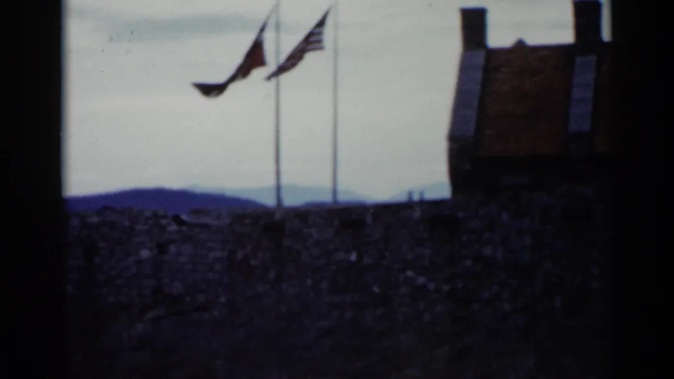 1955 Lake George 8mm frame showing two flags above a stone wall, with hills in the background and a cloudy sky.