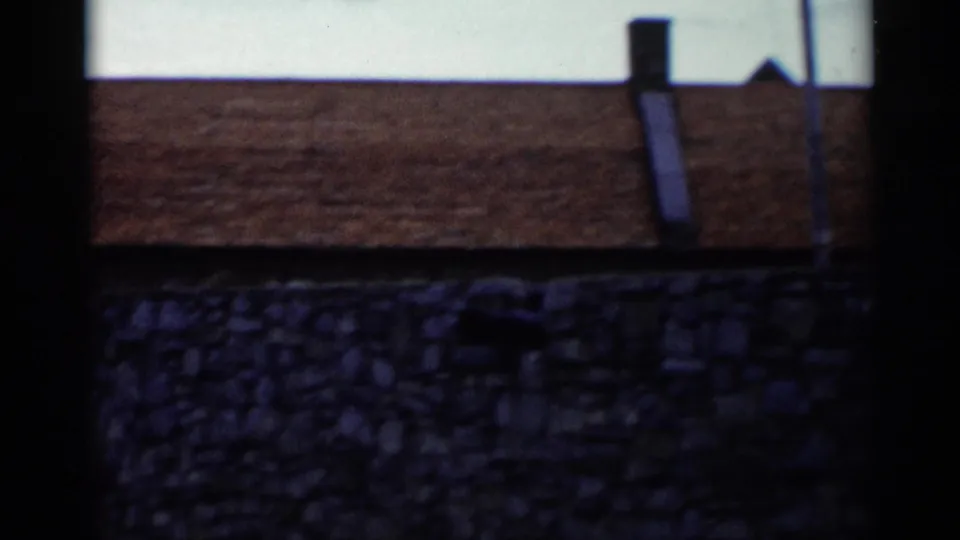 1955 Lake George frame with a roofline or tower behind a stone wall and flagpoles rising into the sky.