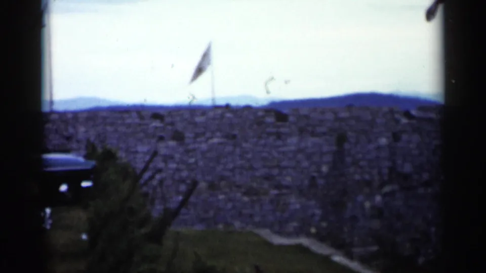 A flagpole rises beyond the stone wall, with hills and a partial dark car visible in this Lake George frame.
