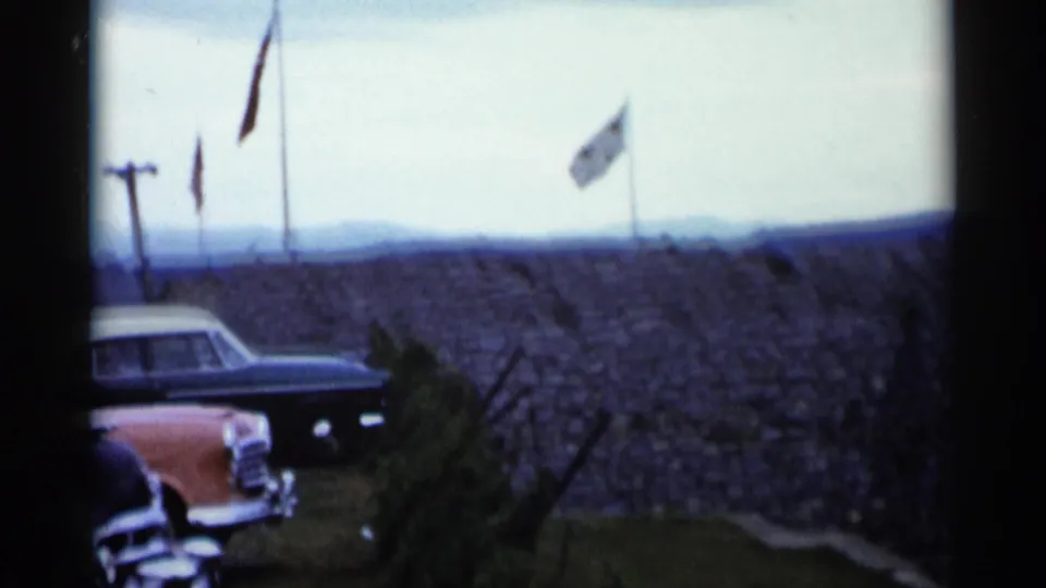 1955 Lake George home-movie frame showing parked cars near a stone overlook wall with flagpoles against a cloudy sky.
