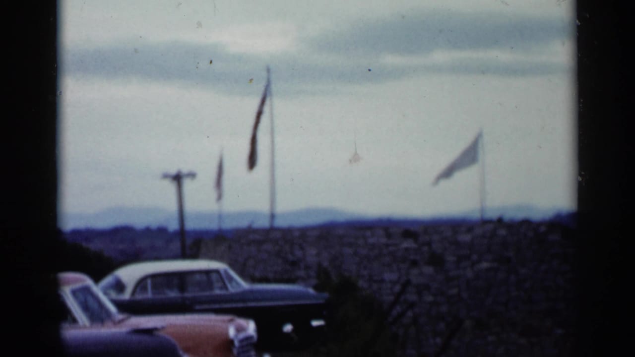 Cars parked near a stone wall with several flags and distant hills behind it.