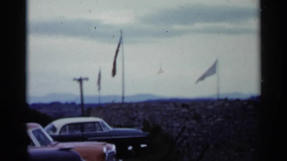 1955 Lake George footage frames parked cars below a stone wall, with flags waving above the overlook and hills in the distance.