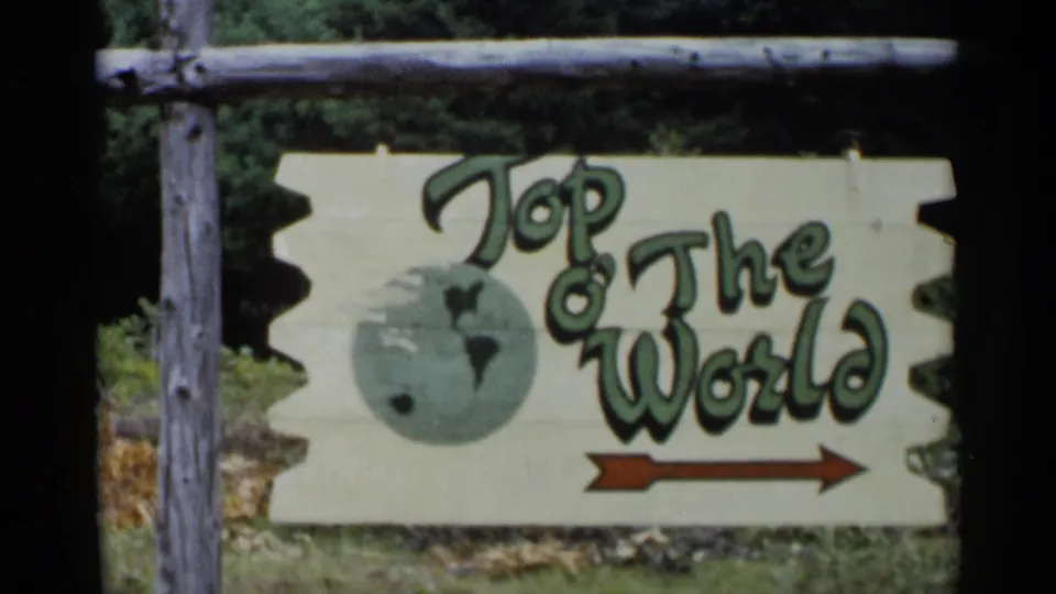 1955 Lake George home-movie frame of a wooden Top o' The World sign with a painted globe and red arrow pointing right.