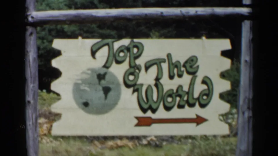 Close 1955 Lake George frame of the Top o' The World wooden sign, including stylized lettering, a globe graphic, and a red arrow.