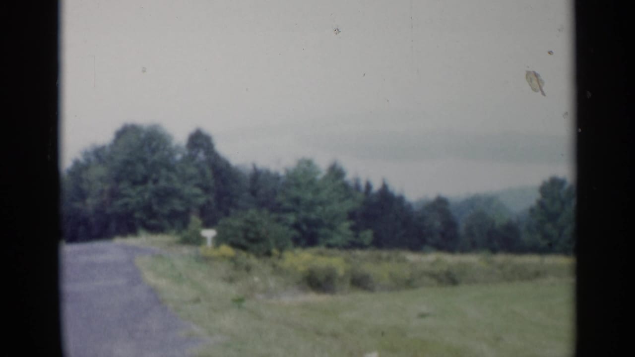1955 Lake George home-movie view of a quiet rural road, open grass, treeline, and distant hazy hills.