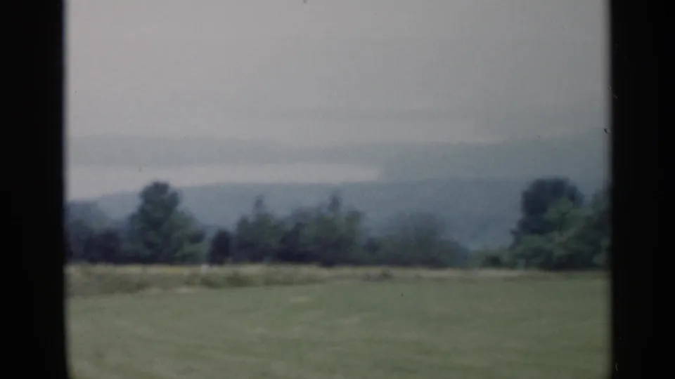 1955 Lake George area countryside view with a grassy foreground, tree line, and misty hills in soft 8mm color.