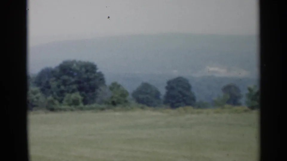 Vintage 8mm countryside frame from Lake George, with a soft grassy slope, tree line, and muted hills beyond.