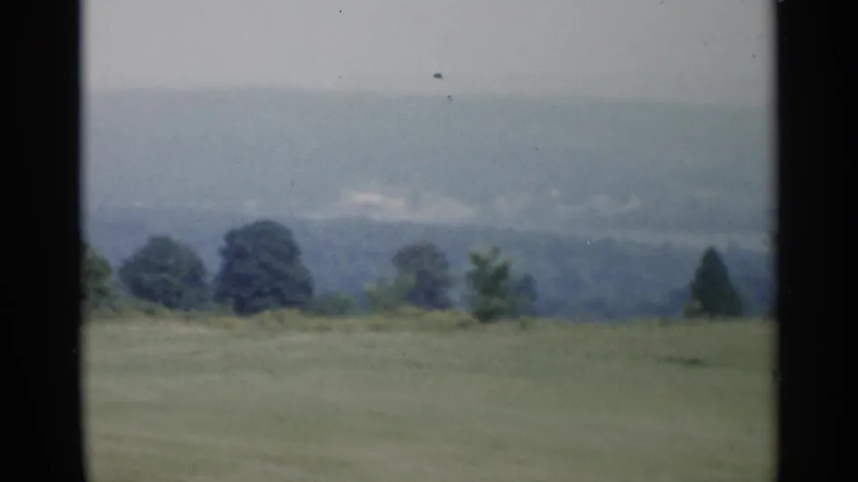 1955 home-movie view near Lake George showing a quiet field, scattered trees, and blue-gray hills under overcast light.