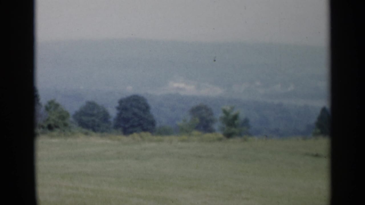 Lake George, New York countryside frame with open grass, dark trees, and hazy hills recorded on 1950s 8mm film.