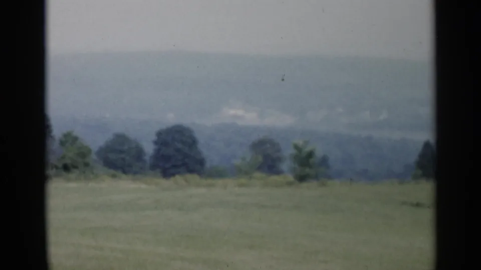 Lake George, New York countryside frame with open grass, dark trees, and hazy hills recorded on 1950s 8mm film.