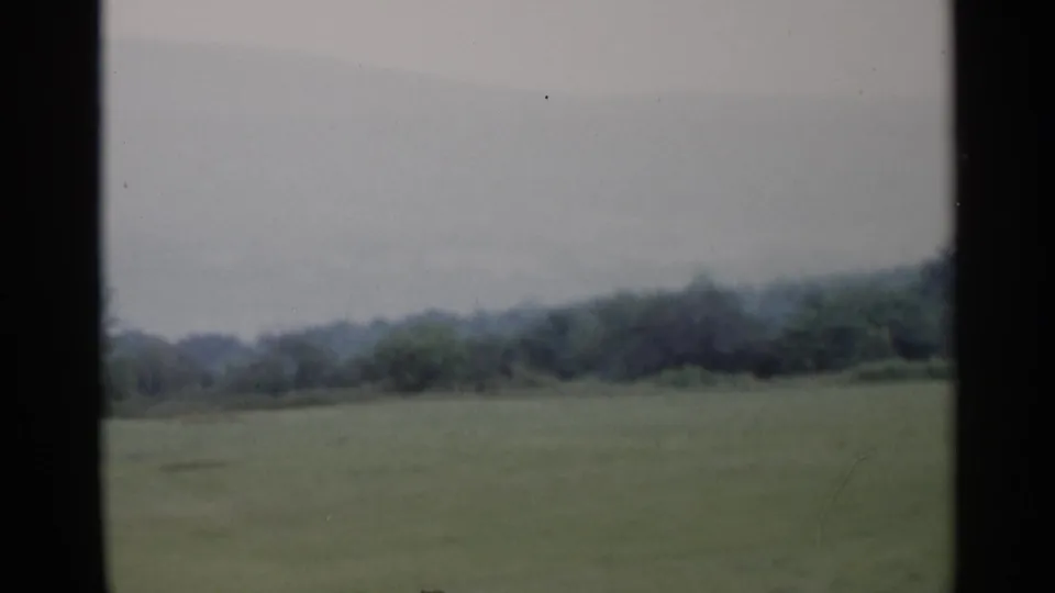 1955 Lake George home-movie frame showing a broad grassy field, distant tree line, and soft hazy hills.
