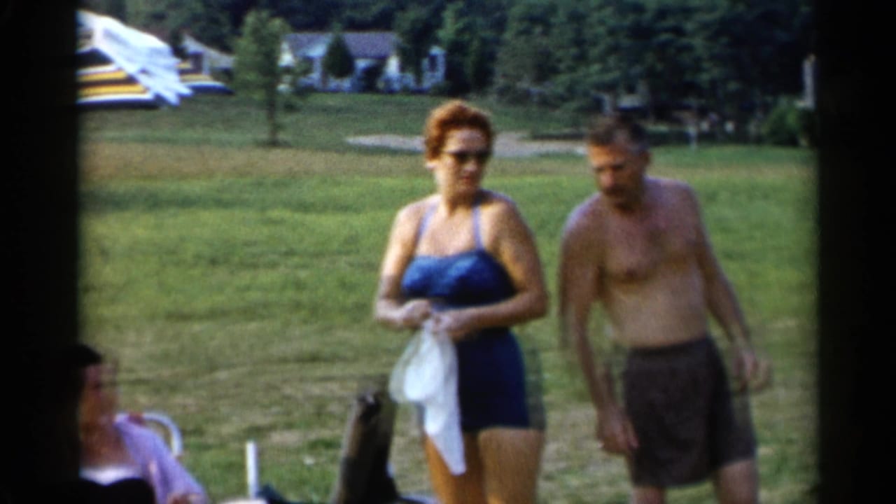 Adults in swimwear stand and sit on grass near a striped umbrella and distant houses.
