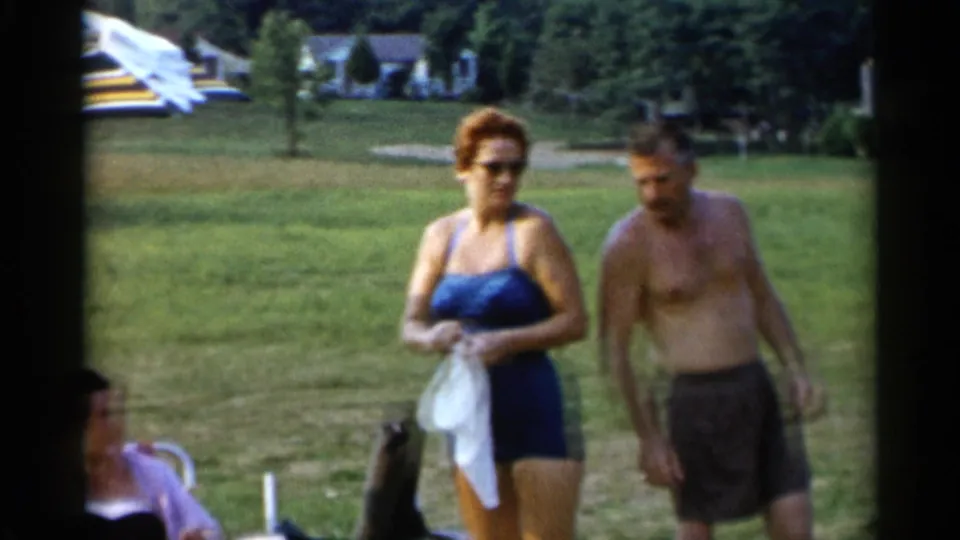 In Lake George, New York, 1955, adults in swimwear gather on grass, with a woman holding a towel and a seated figure partly visible.