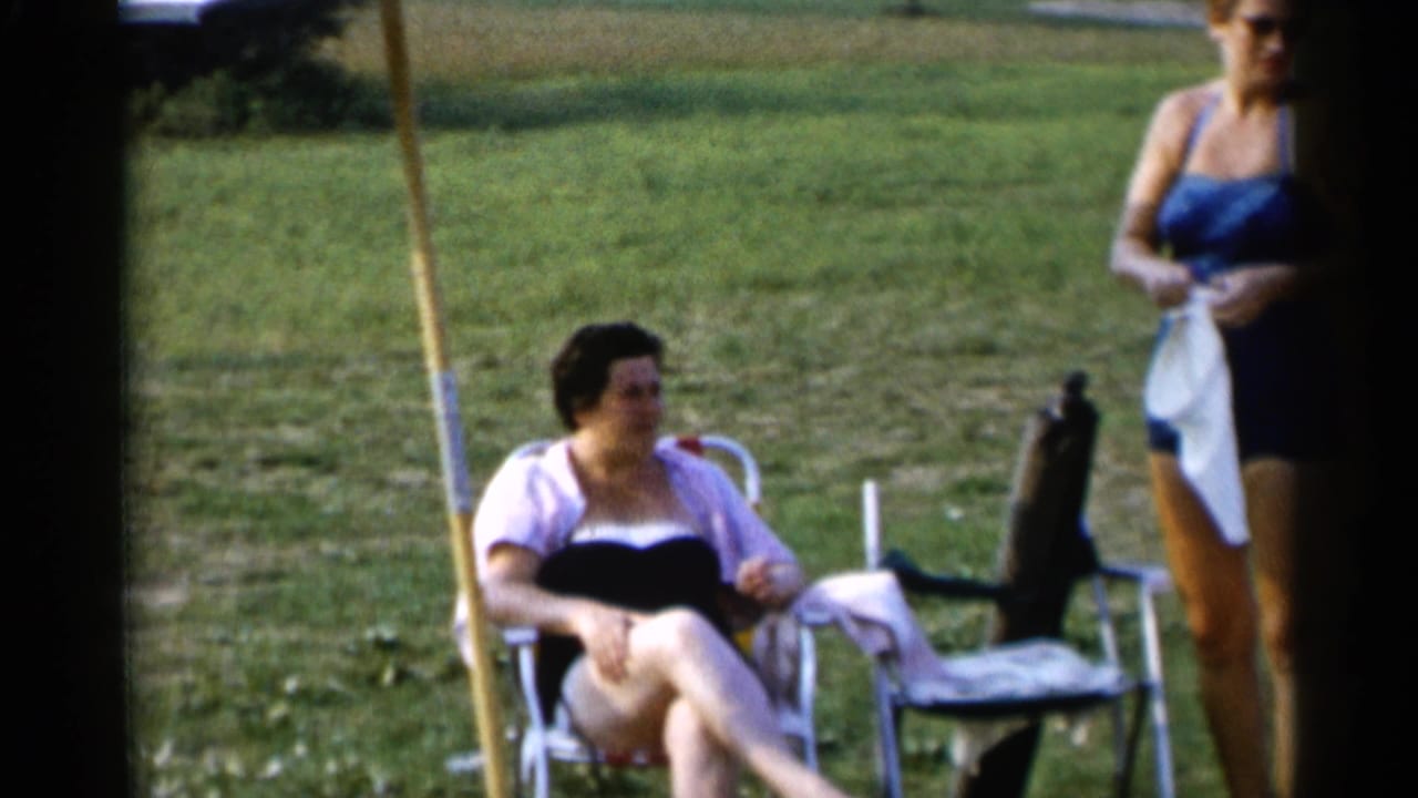 Seated woman in a folding chair and standing woman in a blue swimsuit on a grassy field.