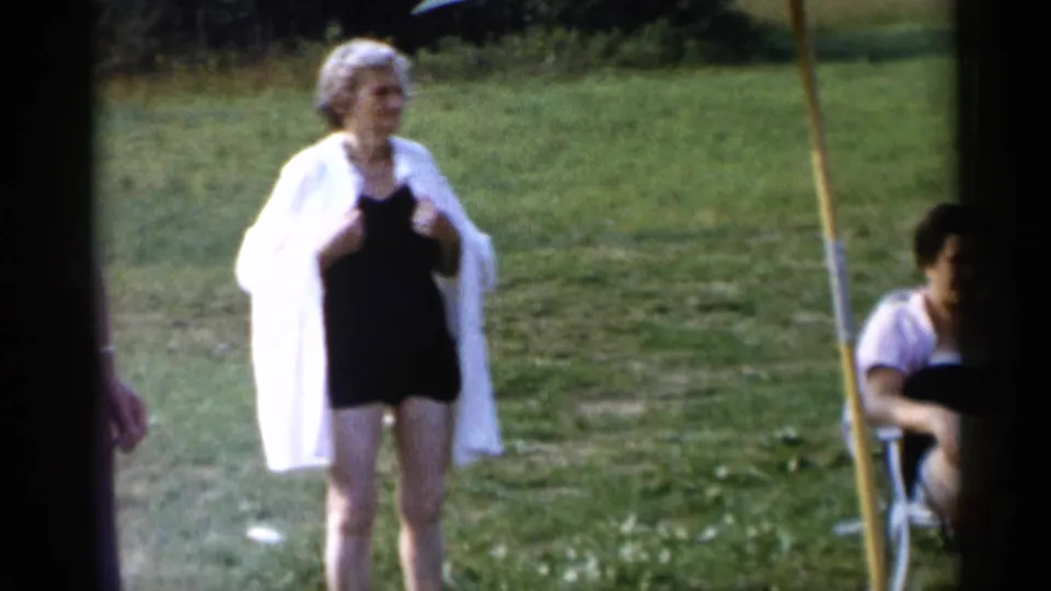 At a Lake George park in 1955, an older woman stands in a black swimsuit and light cover-up while others sit and stand nearby.