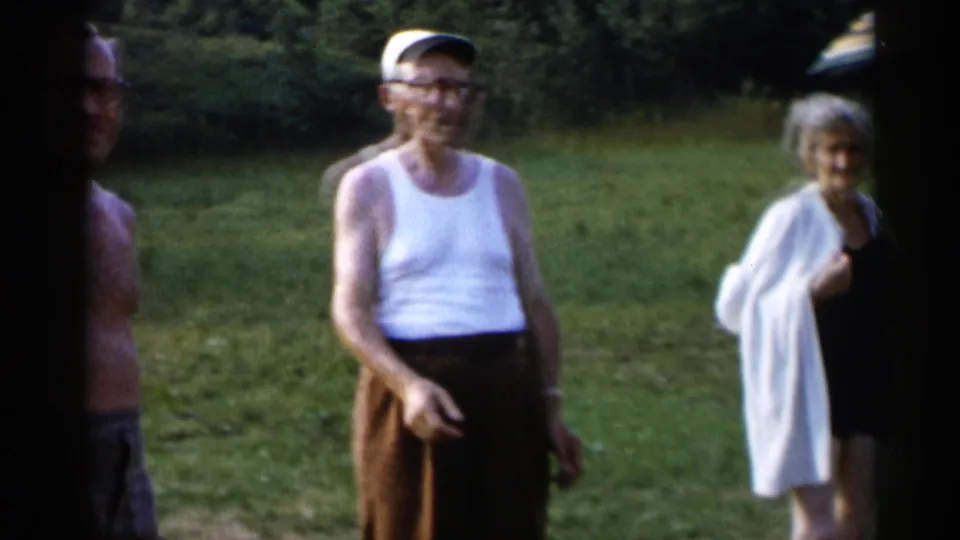The 1955 Lake George home movie shows an older man in a white tank top and cap smiling near an older woman wrapped in a light cover-up.