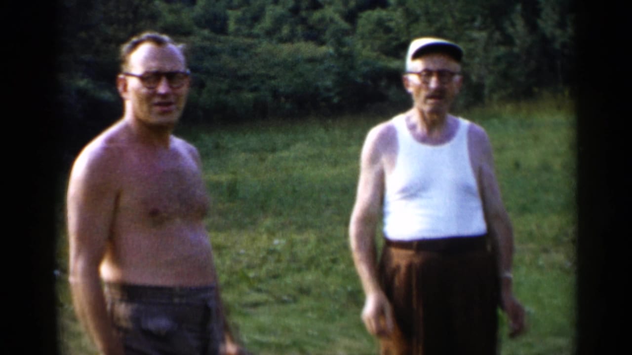 At Lake George in 1955, two adult men stand in a grassy outdoor area, one shirtless and wearing glasses and the other in a white tank top, brown trousers, and a light cap.
