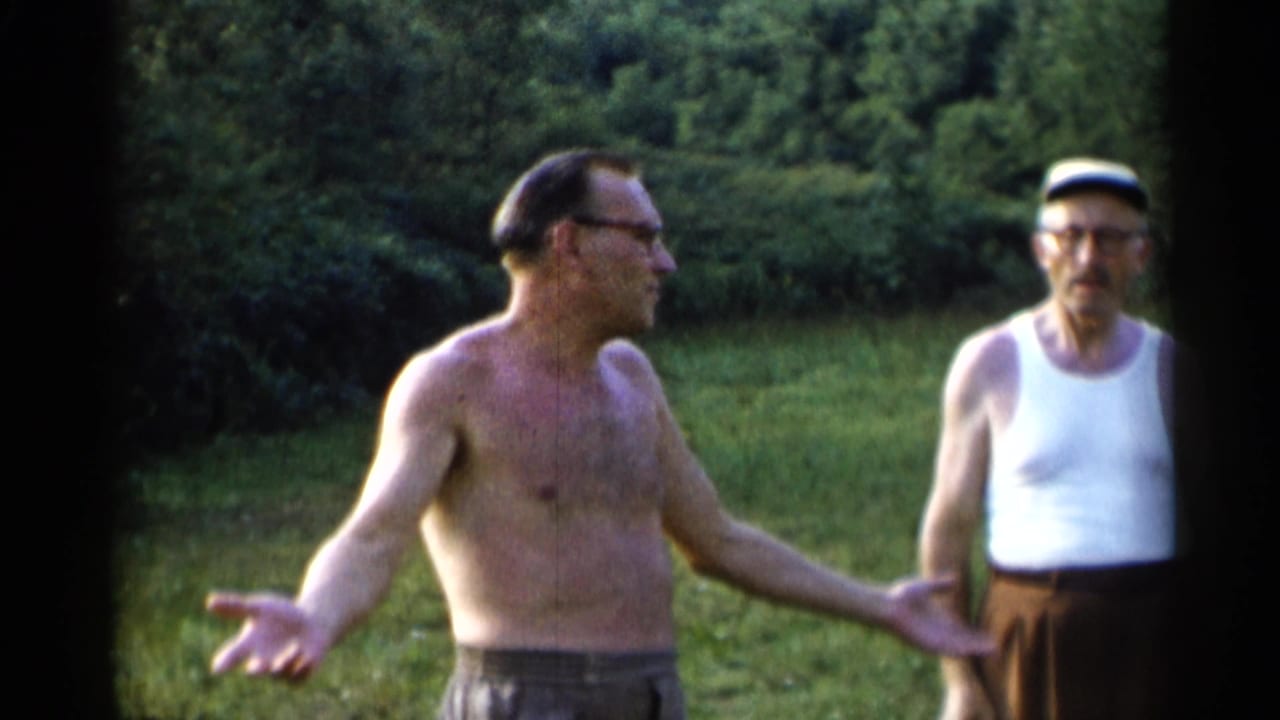 At Lake George in 1955, a shirtless man with glasses gestures toward the camera while a second man in a white tank top and cap stands beside him in a grassy clearing.