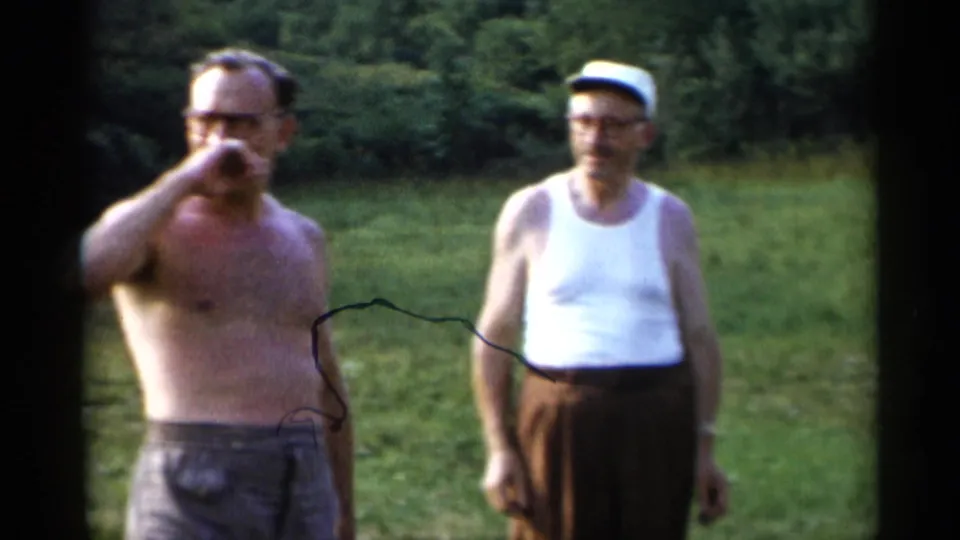 In this 1955 Lake George home-movie frame, a shirtless man with glasses raises a hand near his face as another man in a white tank top and cap looks on from the lawn.