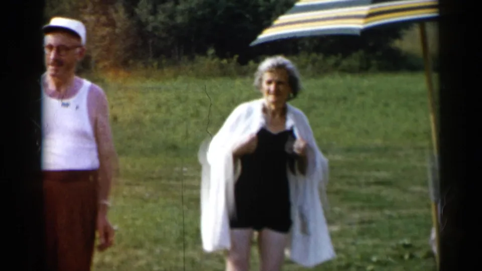 At Lake George in 1955, a woman in a black swimsuit with a white cover-up walks across the grass near a striped umbrella as a man in a white tank top stands to the side.