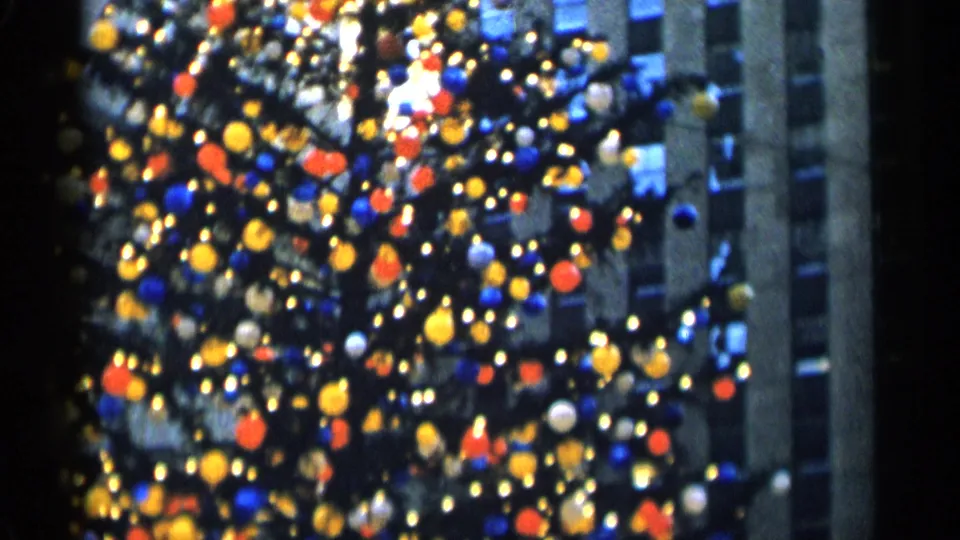 New York City holiday tree in soft 8mm focus, with clustered colored lights, ornaments, and a shadowed building behind it.