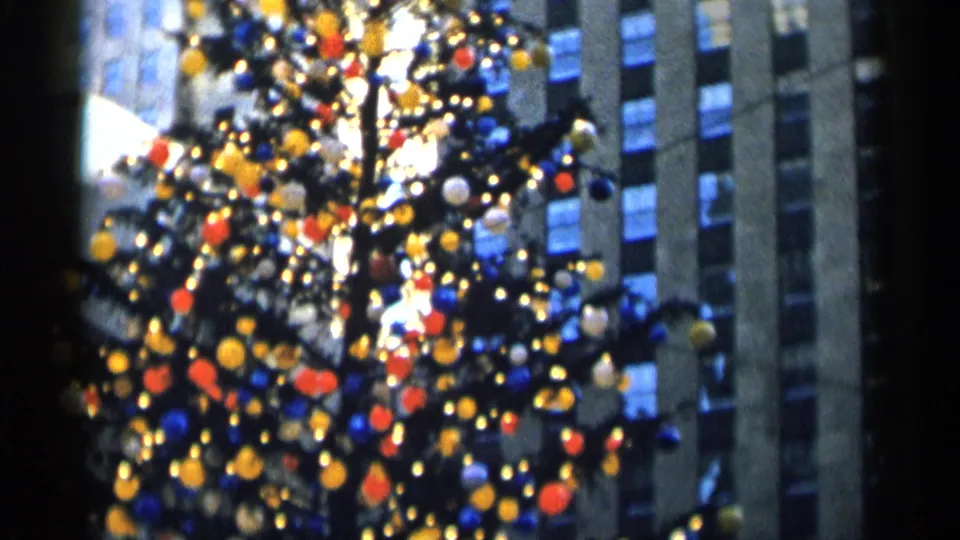 Decorated holiday tree in soft focus, with red, yellow, blue, and white ornaments near dark high-rise architecture.